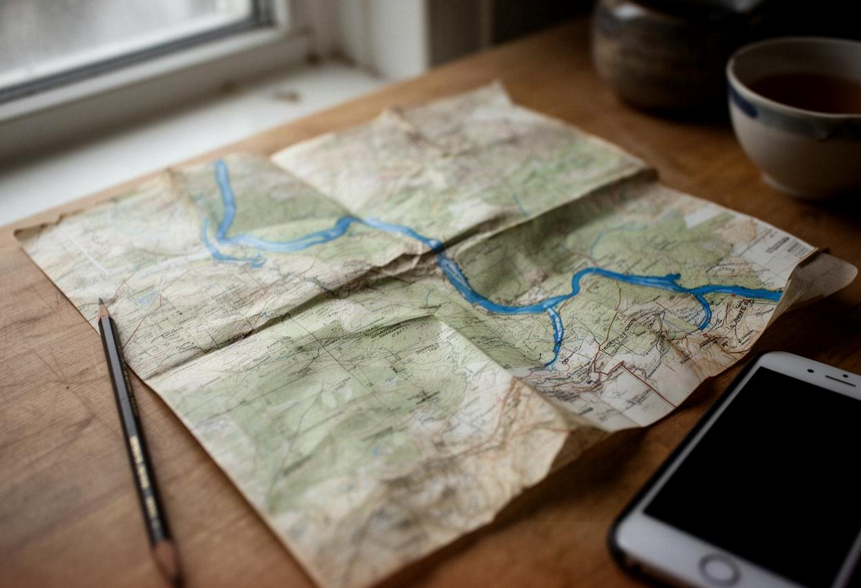 Złożona papierowa mapa z niebieską linią rzeki na drewnianym stole, obok ołówek i telefon